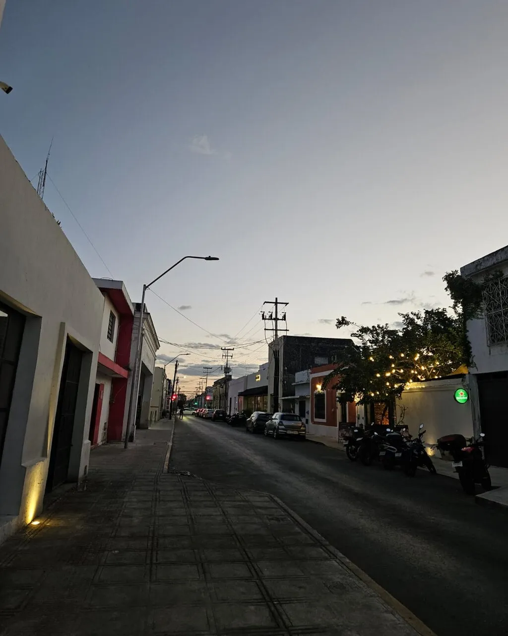 A quiet Mérida street at dusk, with low façades, string lights and a wide stone sidewalk.