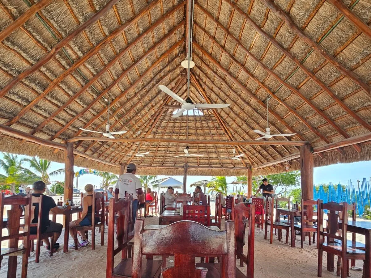 A beachside restaurant under a high thatched palapa roof, with wooden tables set on the sand.